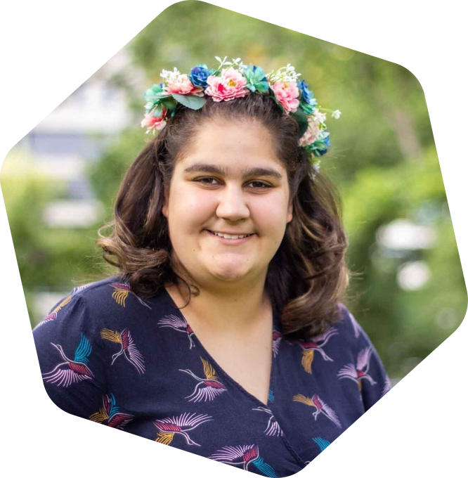 Fel smiling. She is outside amongst greenery and is wearing a flower crown.